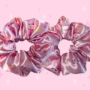 May include: Two pink satin scrunchies with a repeating pattern of cartoon characters and floral designs. The fabric has a shiny, reflective quality. The background is a solid pink colour with white star accents.