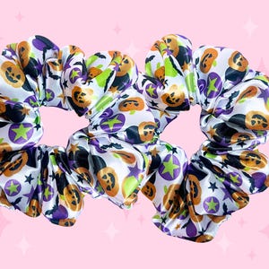 May include: Two Halloween-themed scrunchies. The white satin fabric is patterned with orange jack-o'-lanterns, purple stars, green accents, and black bats. These hair accessories are perfect for adding a festive touch to any hairstyle.