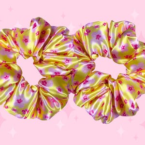 May include: Two hair scrunchies made of a shiny yellow fabric featuring a repeating pattern of small pink flowers and soft purple cloud-like shapes. The scrunchies have a gathered, ruffled texture and a smooth, reflective finish throughout.