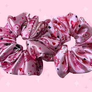 May include: Two pink satin scrunchies with a repeating pattern of teddy bears and hearts. The scrunchies are a light pink color and have a shiny, smooth texture. The background is a matching pink.