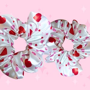 May include: Three white satin scrunchies patterned with red and pink hearts. The scrunchies are set against a pink backdrop with star-shaped sparkles. These hair accessories are ideal for Valentine's Day or general use.
