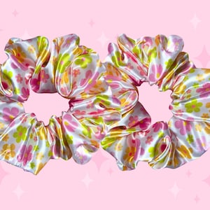 May include: Two hair scrunchies made of a white satin-like fabric featuring a repeating pattern of small five-petaled flowers in shades of bright pink, lime green, and golden yellow. The fabric is gathered into soft, voluminous ruffles.