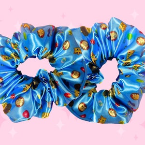 May include: Two bright blue satin scrunchies featuring a repeating pattern of golden crowns, small circular portraits of dogs, and red heart shapes. The fabric has a smooth, reflective sheen and is gathered into a ruffled, circular hair accessory shape.
