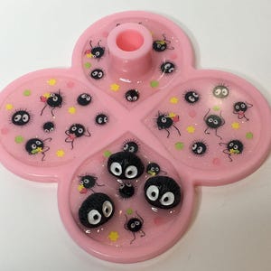 May include: A pink, four-leaf clover-shaped tray with a central cylindrical holder. The tray is decorated with glitter and cartoon soot sprites. The soot sprites are black with large white eyes and are in various poses.