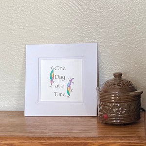 Op de afbeelding: Een wit ingelijst afdruk met de tekst "One Day at a Time" in kleurrijke cursieve letters. De afdruk bevindt zich op een houten oppervlak naast een bruine keramische olieverdamper.