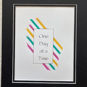 Puede incluir: Una impresión enmarcada en blanco con una cita motivacional, "One Day at a Time", escrita en tinta negra. La cita está rodeada de líneas diagonales de colores en amarillo, rosa y turquesa.