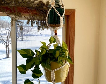 May include: A white macrame plant hanger with two pots, one green and one yellow, hanging from a window. The window is looking out onto a snowy landscape.