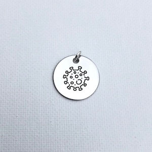 May include: A round, silver-colored pendant with a small loop at the top. The pendant features a black line drawing of a virus. The background is a white, textured surface.