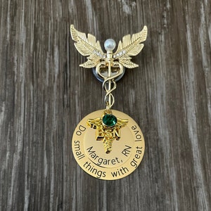 May include: Gold-tone medical caduceus charm with a green gem and a round gold-tone disc with the text "Do small things with great love Margaret, RN".