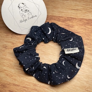 May include: A dark blue scrunchie with a white moon and star pattern. The scrunchie has a white label with the text "Abyss" on it.