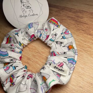 May include: White scrunchie featuring a repeating pattern of llamas in colourful outfits. The scrunchie has a small tag that reads "Abrlyss Creations".