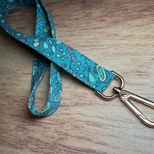May include: A teal fabric lanyard with a gold-coloured metal clasp. The lanyard features a repeating pattern of light blue, green, and pink shapes. The lanyard is lying on a wooden surface.