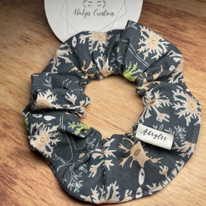 May include: A dark grey scrunchie featuring a floral design in beige and green. The fabric scrunchie has a small white label with the brand name "Abryss Creations".