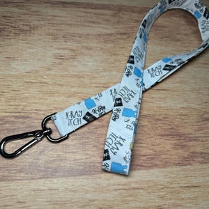May include: A white lanyard with a silver metal clasp. The lanyard is printed with the words "X-RAY TECH" in black and blue, along with related graphics. The lanyard is lying on a wooden surface.