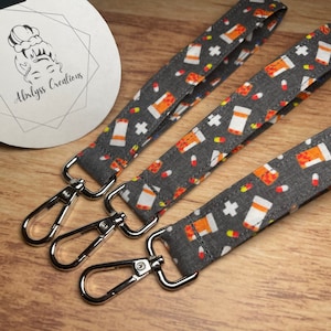 May include: Three fabric wristlet keychains featuring a dark gray background with a repeating pattern of orange and white prescription pill bottles, red and yellow capsules, and white crosses, each finished with a silver metal lobster clasp.