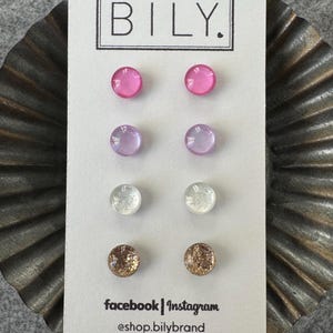 May include: A set of six pairs of stud earrings on a white card with the text "BILY." The earrings are in four different colors: pink, lavender, white, and gold. The card also includes the text "facebook | instagram @shop.bilybrand".