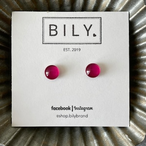 May include: A pair of magenta-colored stud earrings on a white card with the brand name "BILY." and the text "EST. 2019." The card also includes social media information. The earrings are round and have a glossy finish.