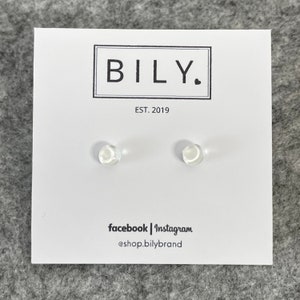 May include: A pair of clear, round stud earrings on a white card with the brand name "BILY." in a black rectangular frame. The card also includes the text "EST. 2019" and social media handles.