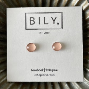 May include: A pair of blush-colored stud earrings on a white card. The card features the brand name "BILY." in a black rectangular box, with "EST. 2019" below. Social media handles are listed at the bottom.