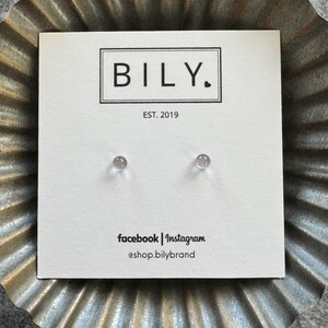 May include: A pair of clear, round stud earrings displayed on a white card. The card features the brand name "BILY" in a black rectangular frame, with "EST. 2019" below. Social media handles are listed at the bottom.