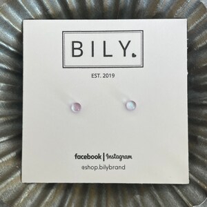 May include: A pair of small, iridescent stud earrings on a white card with the brand name "BILY." in a black box, and the text "EST. 2019." The card also displays social media handles. The earrings are set against a textured, metallic background.