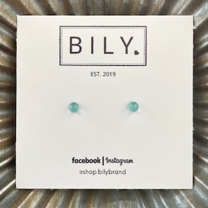 May include: A pair of small, round, light blue stud earrings on a white card. The card has the brand name "BILY." in a black rectangle, with "EST. 2019" below. Social media handles are listed at the bottom.
