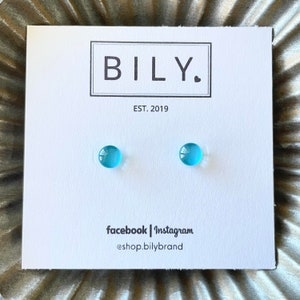 May include: A pair of light blue stud earrings on a white card with the text "BILY." and "EST. 2019". The card also includes social media information: "facebook | Instagram @shop.bilybrand". The earrings are small, round, and a vibrant blue color.