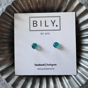 May include: A pair of small, round, translucent teal stud earrings mounted on a white display card. The card features the text BILY. EST. 2019 at the top and facebook | Instagram @shop.bilybrand at the bottom in black sans-serif font.