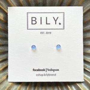 May include: A pair of light blue stud earrings on a white card with the brand name "BILY." and the text "EST. 2019." The card also includes social media handles. The earrings are small, round, and a soft blue color.