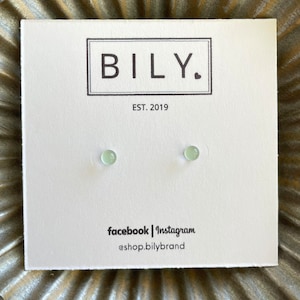 May include: A pair of small, pale green stud earrings on a white card. The card features the brand name "BILY." in a black rectangular box, with "EST. 2019" below. Social media handles are listed at the bottom.