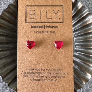 May include: A pair of small, translucent red heart-shaped stud earrings attached to a rectangular brown cardboard display card. The card features the text BILY, social media handles, and a donation message for a local pet rescue.