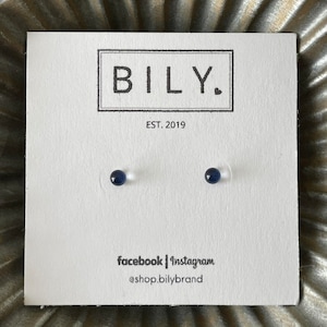 May include: A pair of small, dark blue stud earrings on a white card. The card has the brand name "BILY." in a black rectangular box, with "EST. 2019" below. Social media handles are listed at the bottom.