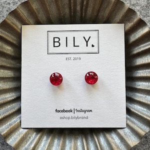 May include: A pair of small, round, red stud earrings on a white card with the brand name "BILY." and the text "EST. 2019." The card also includes social media information. The earrings are set against a textured, metallic background.