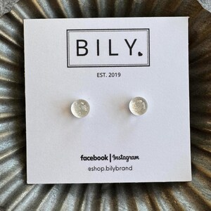 May include: A white card with the text "BILY. EST. 2019" and the social media handles "facebook | instagram @shop.bilybrand". Two small, clear, round, sparkly earrings are displayed on the card.
