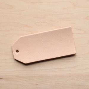 May include: A light pink leather tag with a hole punched in the top. The tag is on a light wood surface.