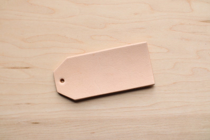 May include: A light pink leather tag with a hole punched in the top. The tag is on a light wood surface.