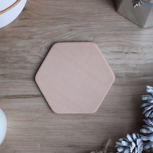 May include: A hexagonal, light pink leather coaster sits on a wooden surface. The coaster is surrounded by decorative elements, including a white ceramic pot, a silver metal object, and white-painted pinecones. The coaster is a neutral color.