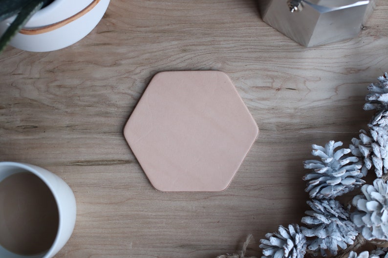 May include: A hexagonal, light pink leather coaster sits on a wooden surface. The coaster is surrounded by decorative elements, including a white ceramic pot, a silver metal object, and white-painted pinecones. The coaster is a neutral color.