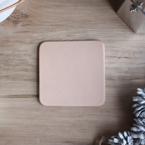 May include: A square, light pink leather coaster sits on a wooden surface. The coaster is surrounded by a white mug, a silver coffee maker, and white-painted pinecones. The scene is shot from above, creating a flat lay composition.
