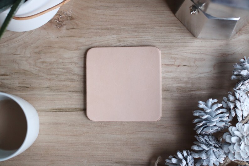 May include: A square, light pink leather coaster sits on a wooden surface. The coaster is surrounded by a white mug, a silver coffee maker, and white-painted pinecones. The scene is shot from above, creating a flat lay composition.