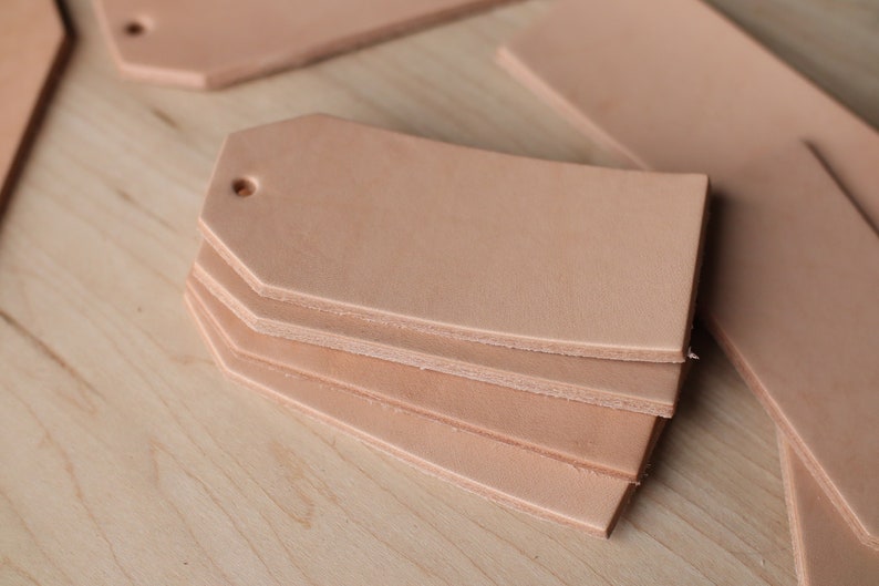 May include: A stack of four unfinished leather tags with rounded corners and a hole punched in the top. The tags are a light brown color and are arranged on a light wood surface.