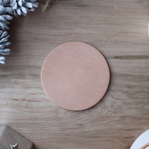 May include: A round, light brown leather coaster sits on a wooden surface. The coaster is surrounded by decorative elements, including white pinecones and a white ceramic mug. The natural wood grain provides a warm backdrop.