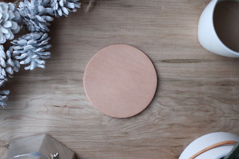 May include: A round, light brown leather coaster sits on a wooden surface. The coaster is surrounded by decorative elements, including white pinecones and a white ceramic mug. The natural wood grain provides a warm backdrop.