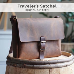 Può includere: Una borsa a tracolla in pelle marrone con un'ampia patta frontale e un singolo cinturino verticale fissato da una fibbia in metallo. Il testo Traveler's Satchel DIGITAL PATTERN è visualizzato in alto. La borsa ha una finitura liscia e opaca.