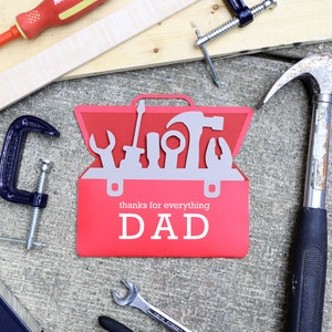 May include: A red toolbox-shaped card with the words "thanks for everything DAD" is surrounded by tools like a hammer, screwdrivers, and a clamp. A wooden plank and a level are also visible, suggesting a Father's Day or handyman theme.