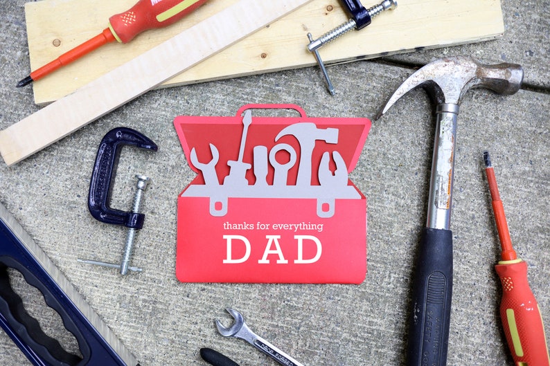 May include: A red toolbox-shaped card with the words "thanks for everything DAD" is surrounded by tools like a hammer, screwdrivers, and a clamp. A wooden plank and a level are also visible, suggesting a Father's Day or handyman theme.
