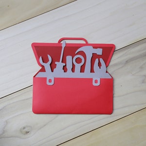 May include: A red paper toolbox with a handle and silver tools inside. The tools include a wrench, screwdriver, hammer, and pliers. The toolbox is set against a light wood grain background.
