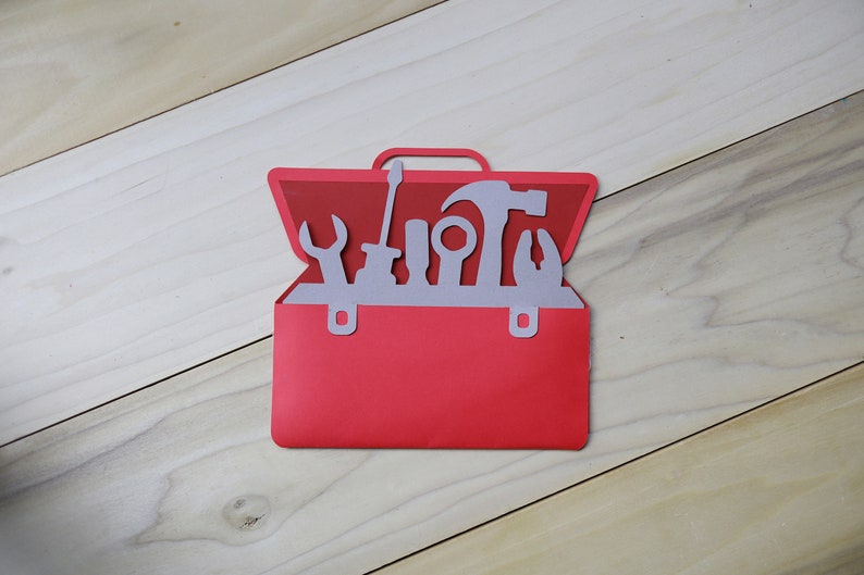 May include: A red paper toolbox with a handle and silver tools inside. The tools include a wrench, screwdriver, hammer, and pliers. The toolbox is set against a light wood grain background.