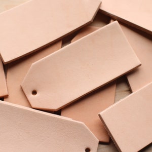 May include: A pile of light brown leather tags with rounded corners and a hole punched in the top. The tags are arranged in a random pattern.