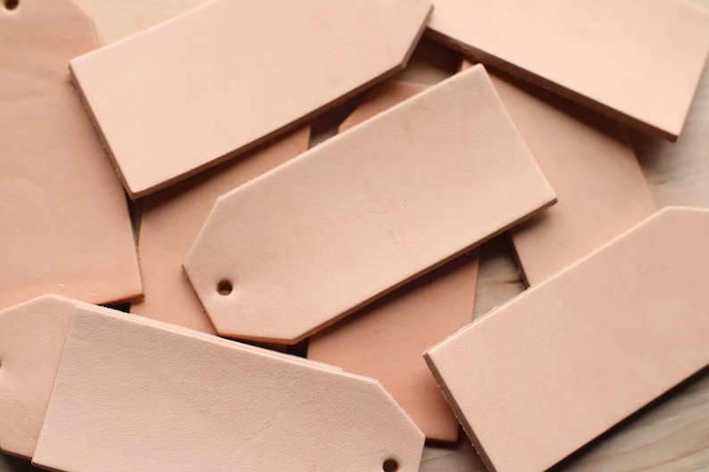 May include: A pile of light brown leather tags with rounded corners and a hole punched in the top. The tags are arranged in a random pattern.